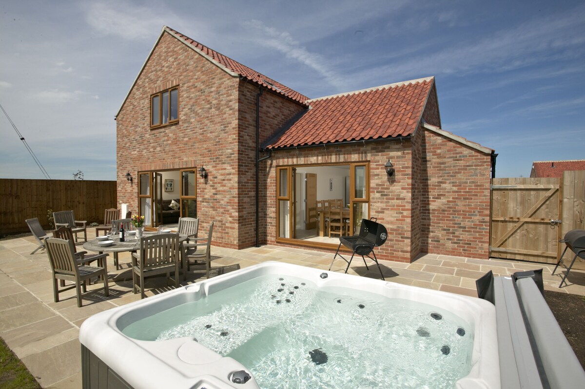 A private hot tub sits on a stone patio in the foreground, while a spacious garden area features wooden dining furniture. The cottage, with its brick exterior and red-tiled roof, is visible in the background, showcasing large glass doors that open onto the patio.