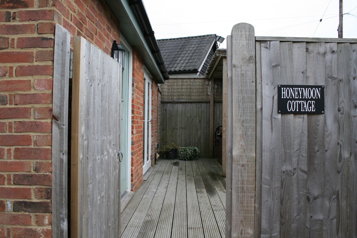 A wooden gate opens to a private deck area leading to the entrance of the Honeymoon Cottage. The exterior features a combination of brick and wood, with a sign identifying the cottage. Potted plants add a touch of greenery to the space.