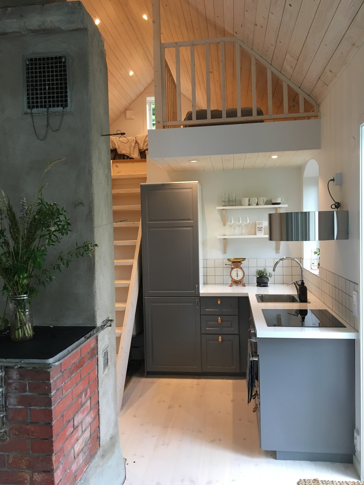 A modern kitchen area is showcased, featuring sleek grey cabinetry and a black countertop. A stainless steel sink is positioned beneath a window. Stairs lead to an upper sleeping loft, with wooden accents present throughout. Natural light filters in, enhancing the inviting atmosphere.