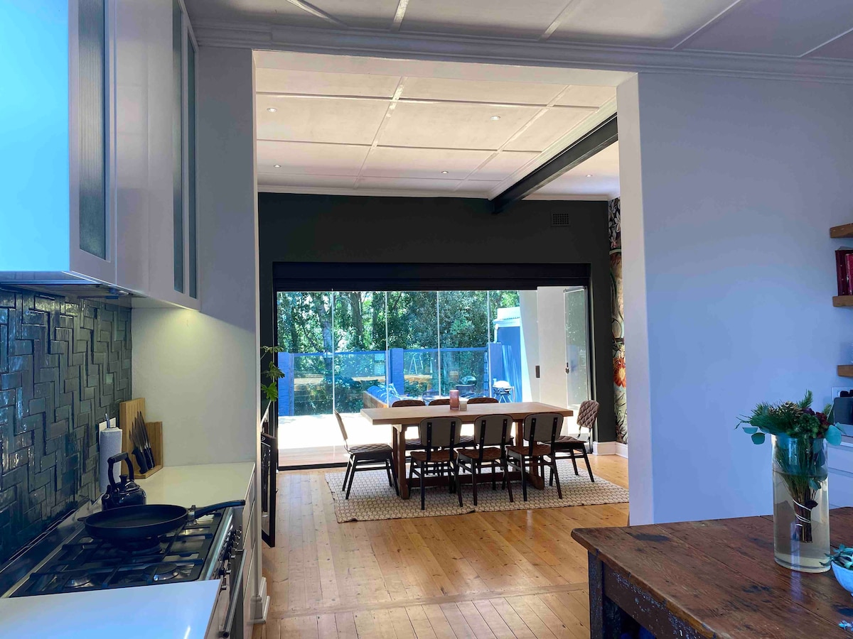 A modern kitchen features sleek cabinetry and high-end appliances, which opens to a spacious dining area. Large glass doors provide a view of the outdoor seating area. Natural light floods the space, highlighting the wooden flooring and minimalist decor.