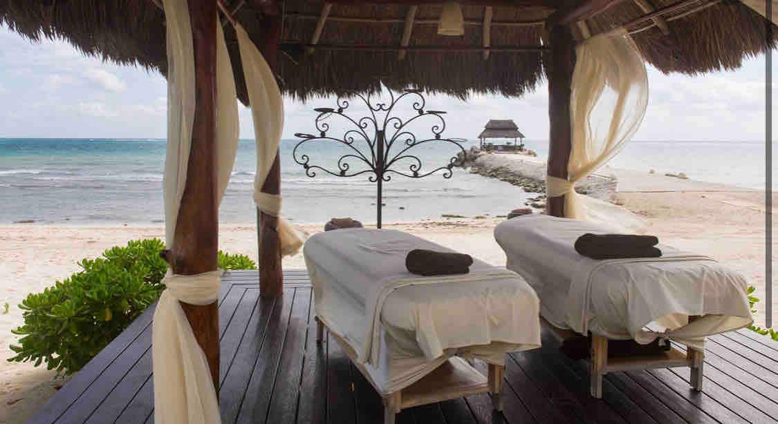 A serene outdoor massage area is observed, featuring two massage tables adorned with rolled towels. The space is shaded by a thatched roof with flowing curtains. A calm sea view extends beyond the wooden deck, inviting relaxation and tranquility.