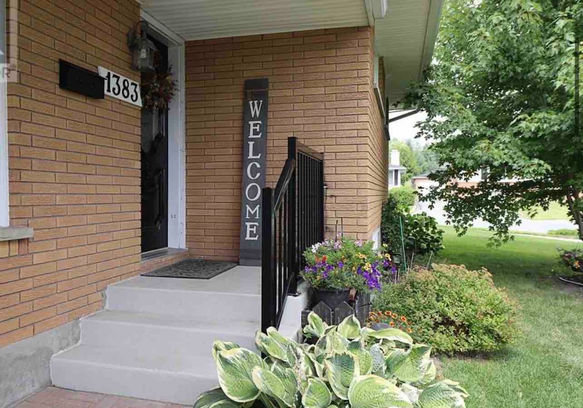 The entrance of the home features a welcoming sign beside the door. Steps lead up to the front door, with a neat welcome mat in place. Lush, green plants and colorful flowers are situated around the porch, enhancing the inviting atmosphere.