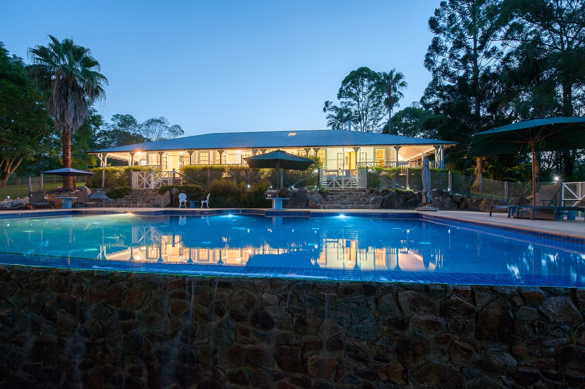 The image captures a tranquil swimming pool illuminated by soft evening light, reflecting the surrounding palms and the cottage. The spacious outdoor area features lounge chairs and umbrellas, with the nearby cottage building showcasing large windows that reveal a warm glow from within.