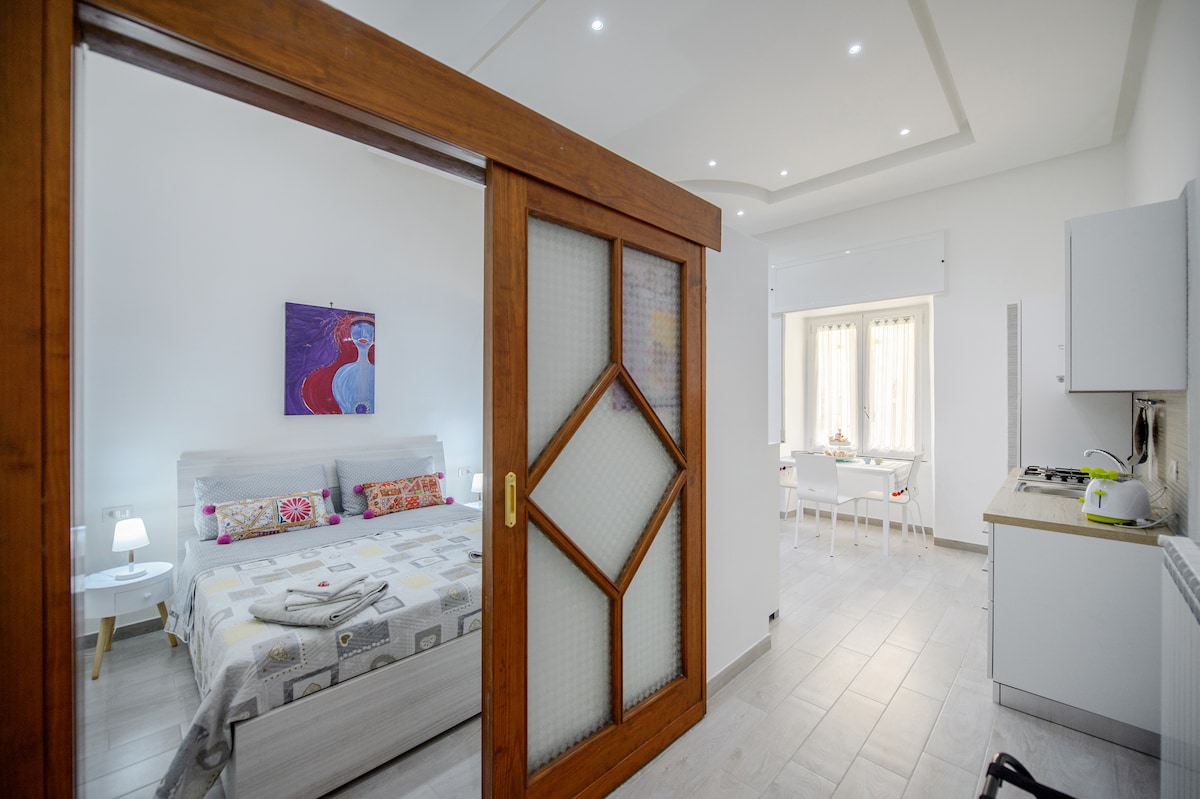 A spacious studio apartment is visible, featuring a cozy sleeping area with a large bed adorned with decorative pillows. A wooden sliding door separates the bedroom from the kitchen and dining area, where a small table is positioned by a window allowing natural light.