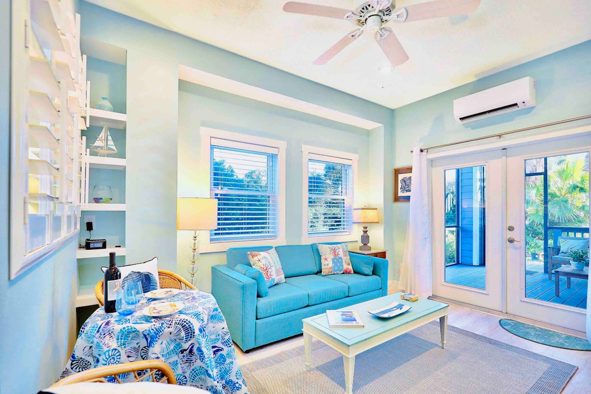 A light-filled living area features a turquoise couch complemented by decorative pillows. A small coffee table is centered in the space, with a patterned tablecloth draping over a rattan side chair. Double glass doors lead to the outdoor deck, framed by light curtains.