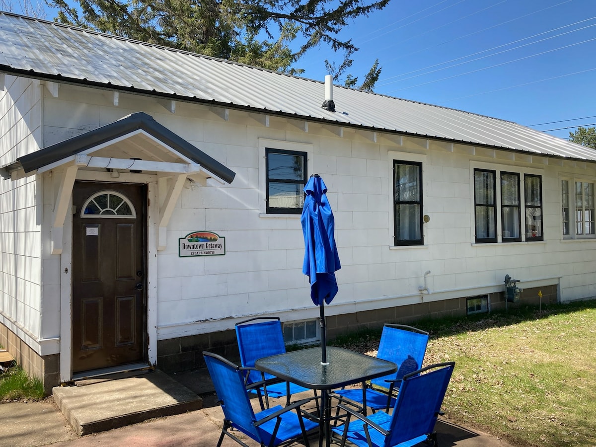 The exterior of Downtown Getaway features a white facade with a metal roof and multiple windows. A small patio area is visible with a blue umbrella over a table and four chairs, offering a spot for outdoor seating. A welcoming front door is centrally located.