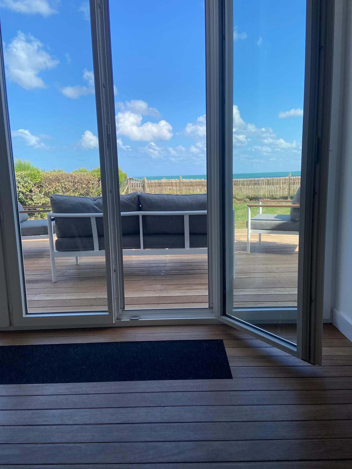 A view from the interior through open glass doors reveals a spacious outdoor terrace. Comfortable seating is arranged on the deck, overlooking lush greenery and the sea under a bright blue sky with soft clouds.