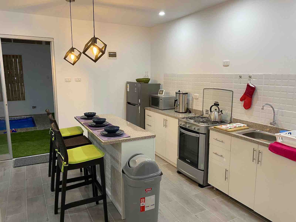 A modern kitchen is equipped with stainless steel appliances, including a refrigerator and gas stove. A breakfast bar with four stools is situated next to a countertop. Soft lighting from pendant lamps enhances the space's functionality. A small entrance leads to an outdoor area visible in the background.