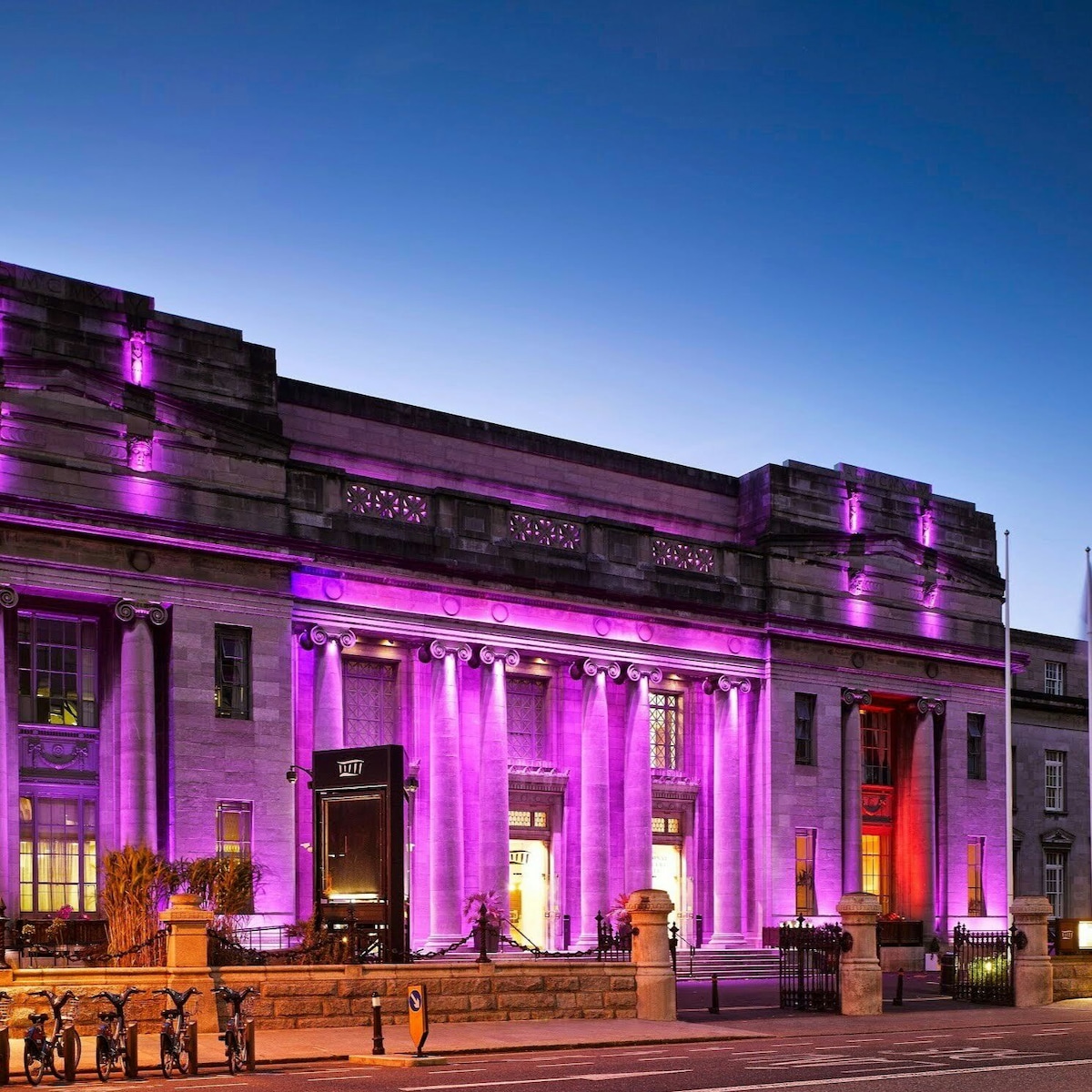 An historic building stands prominently, illuminated by purple and soft orange lighting. Large columns support its sturdy façade, showcasing intricate architectural details. The evening sky transitions into twilight, enhancing the visual appeal of this vibrant city landmark.