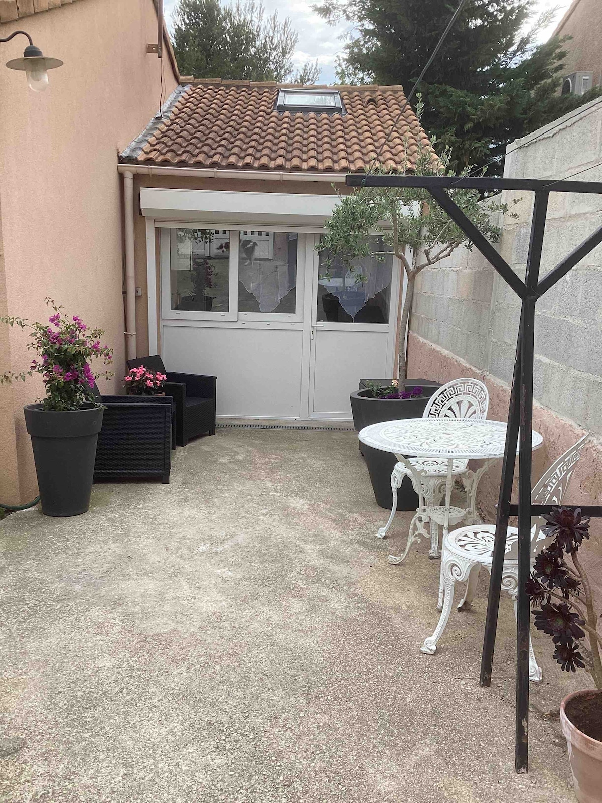 A private outdoor area is seen, featuring a small round table with two decorative chairs. Potted plants with flowers add color, while a clothes drying rack stands in one corner. The space is enclosed by a low stone wall and is accessible through a glass door.