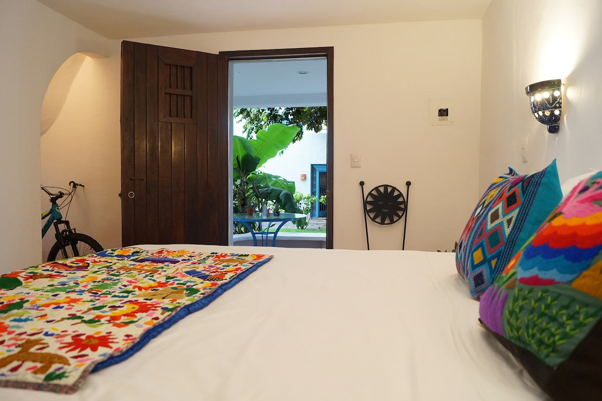 A private room features a spacious bed adorned with a colorful embroidered blanket and decorative pillows. A door leads to a patio view with greenery. A bicycle is parked nearby, and a small table and chair are visible in the outdoor space.
