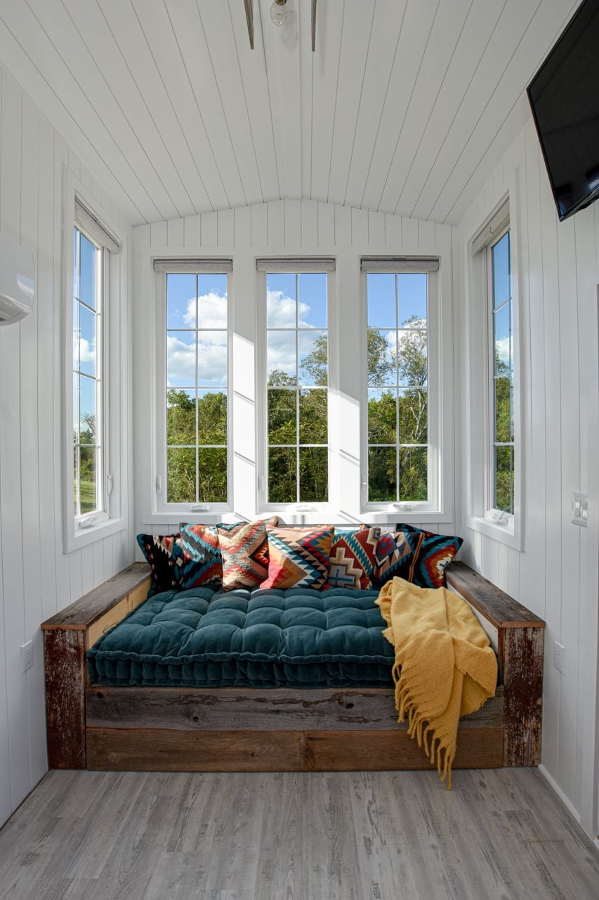 A cozy seating area features a handcrafted daybed framed in rustic wood, adorned with colorful patterned cushions and a soft, yellow throw. Large windows allow plenty of natural light, offering views of the surrounding landscape.