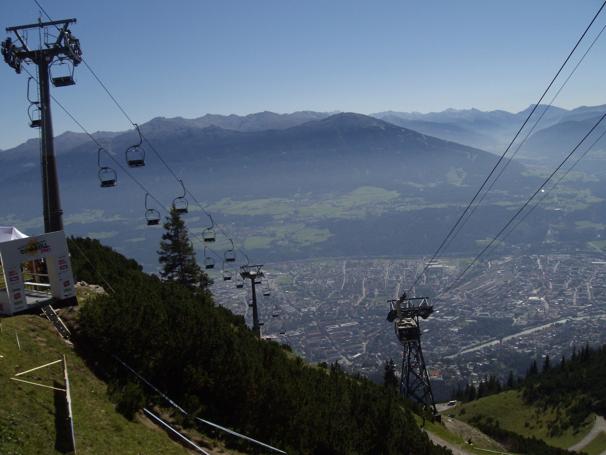 A scenic view captures the city of Innsbruck from a mountain perspective. Several ski lift chairs are visible on the left, ascending through clear blue skies. The valley below showcases the city's layout amid green fields and distant mountains, offering insight into the area's natural beauty.