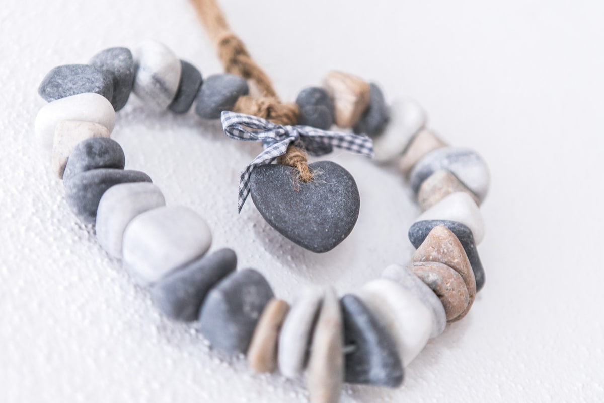 A heart-shaped decoration made from smooth stones in shades of gray, white, and beige is displayed on a textured white background. The arrangement of the stones creates a natural and calming appearance, while a small dark stone in the shape of a heart is suspended with a checkered ribbon.