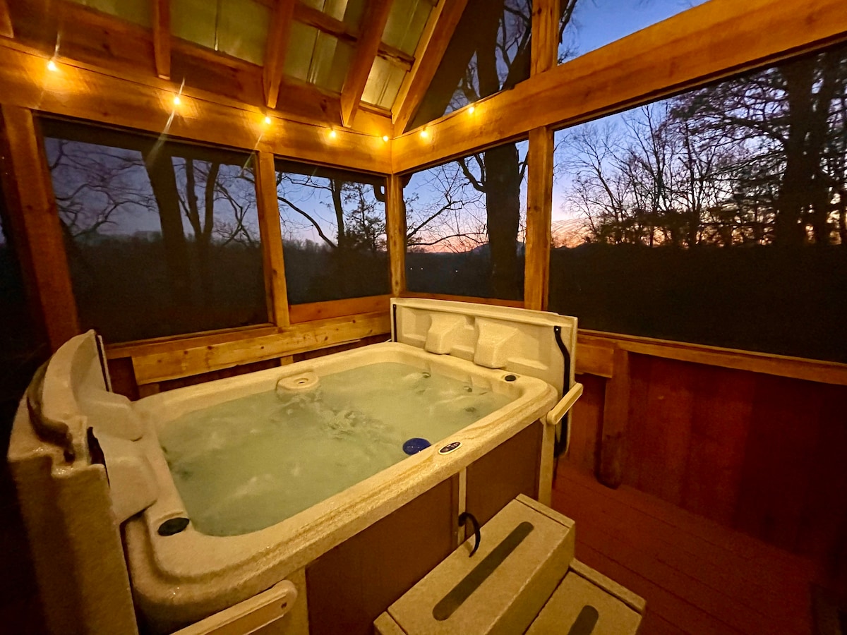 A hot tub is situated in a screened-in area, surrounded by wooden beams and panels. Warm light strings add a cozy touch, while a sunset view can be seen through the trees in the background, enhancing the serene atmosphere.