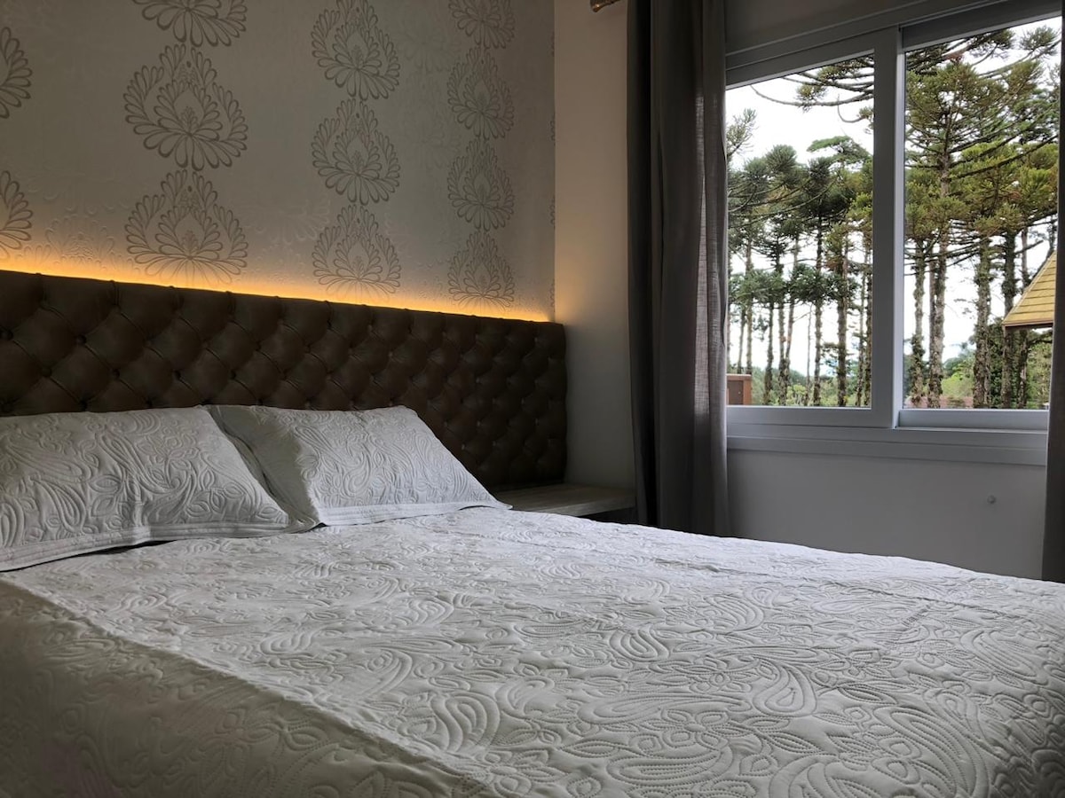 A spacious bed is positioned against a textured wall, featuring elegant lighting above. Natural light filters in through a large window, providing a view of tall trees outside, enhancing the overall calm ambiance of the sleeping area.