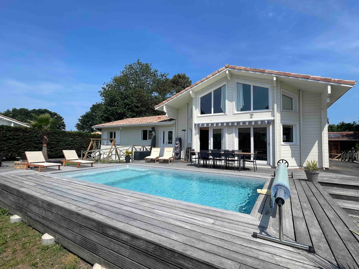 The villa features a heated swimming pool surrounded by a spacious wooden deck. Sun loungers are arranged nearby, and a dining area is visible on the deck. The structure presents large windows and a light exterior, set against a backdrop of greenery.