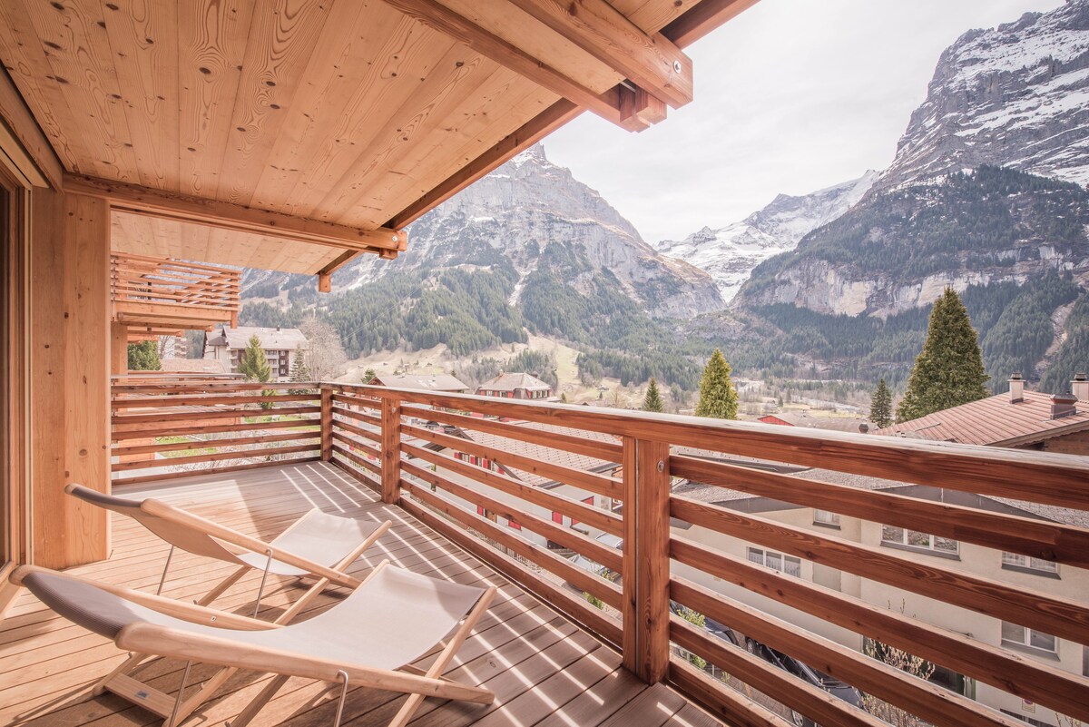 A spacious balcony is presented with wooden decking and slatted railings, providing expansive views of the surrounding mountains. Two lounge chairs are positioned for relaxation while enjoying the impressive scenery, characterized by steep peaks and lush valleys.
