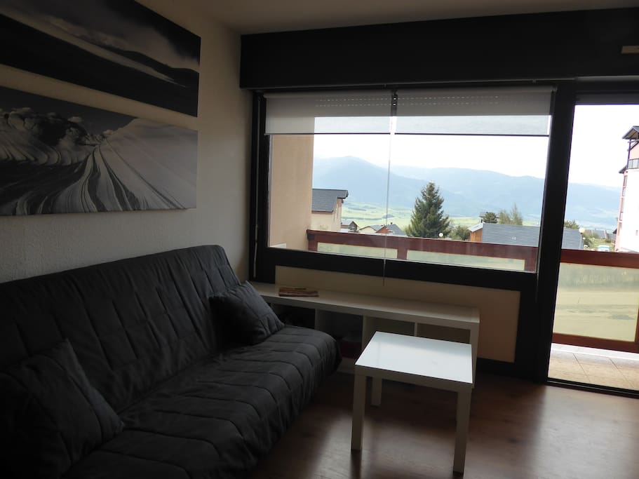 la pièce principale avec le clic-clac, porte ouverte sur le balcon où se trouvent une petite table et 2 chaises