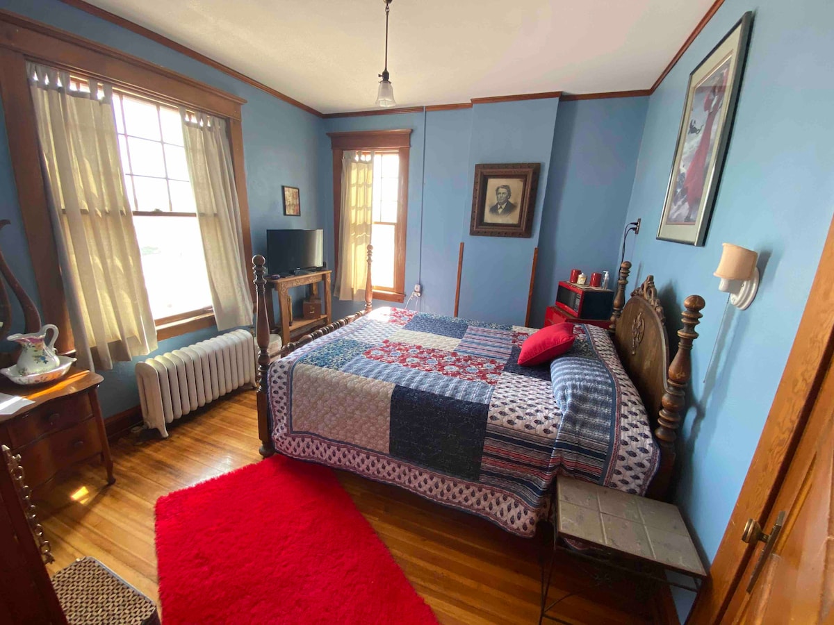 A cozy bedroom features a spacious bed adorned with a colorful patchwork quilt. Natural light enters through large windows, complementing the blue walls. A small television is positioned near the bed, and decorative items are displayed on a nightstand and an accent table.