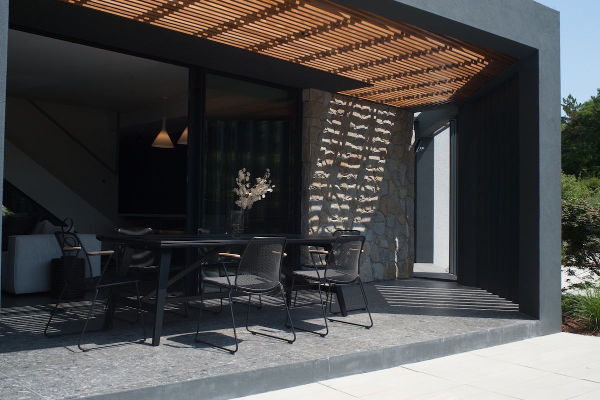 An outdoor dining area is presented, featuring a sleek black table surrounded by several modern chairs. A wood slatted roof provides shade, while a stone wall adds texture. Large glass doors connect this space to the interior, inviting natural light.