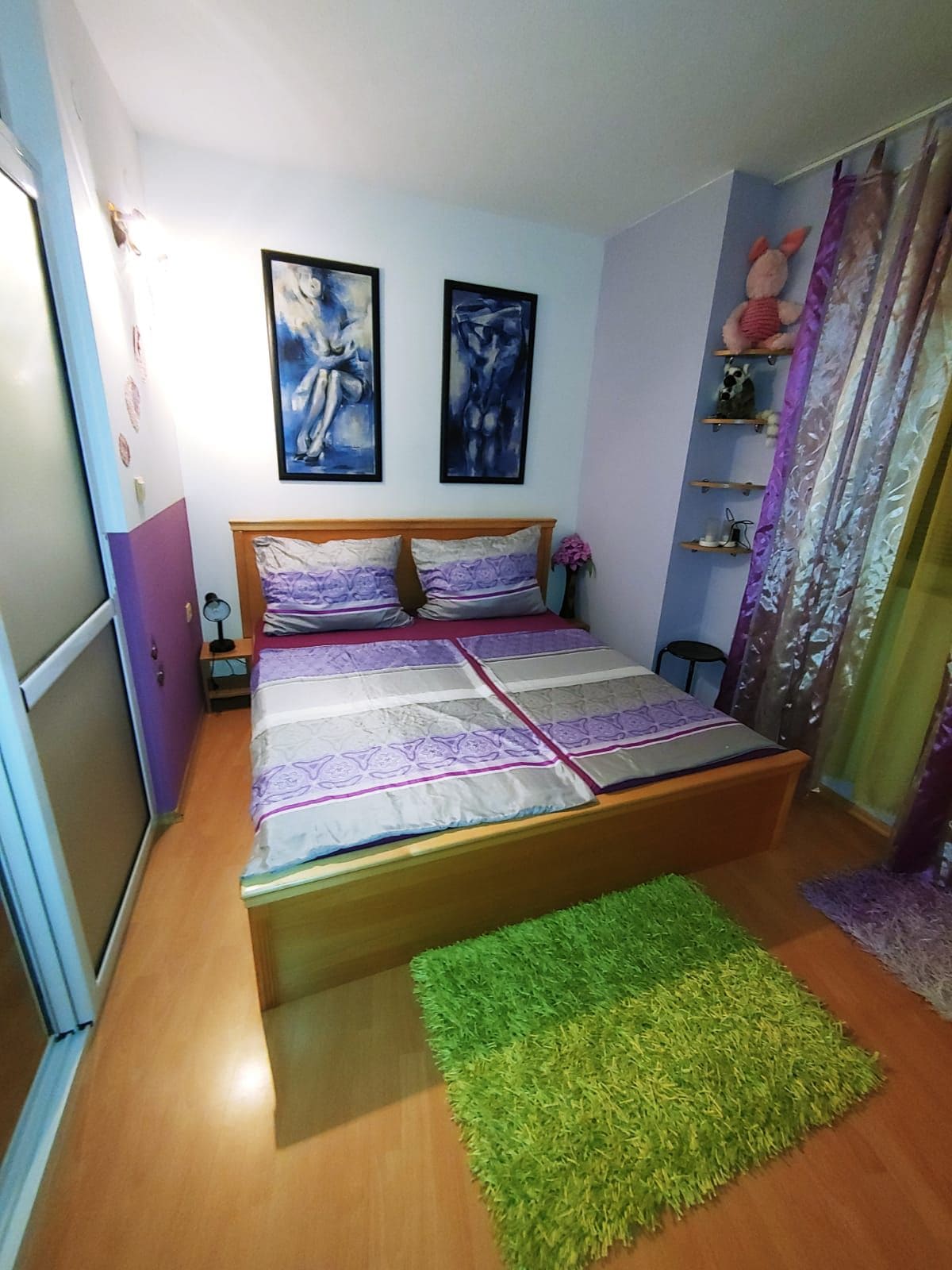 A cozy bedroom is featured with a wooden bed adorned with patterned bedding. Vibrant artwork decorates the walls, while colorful drapes and a plush green rug enhance the space. Natural light streams in through a glass door, creating an inviting ambiance.