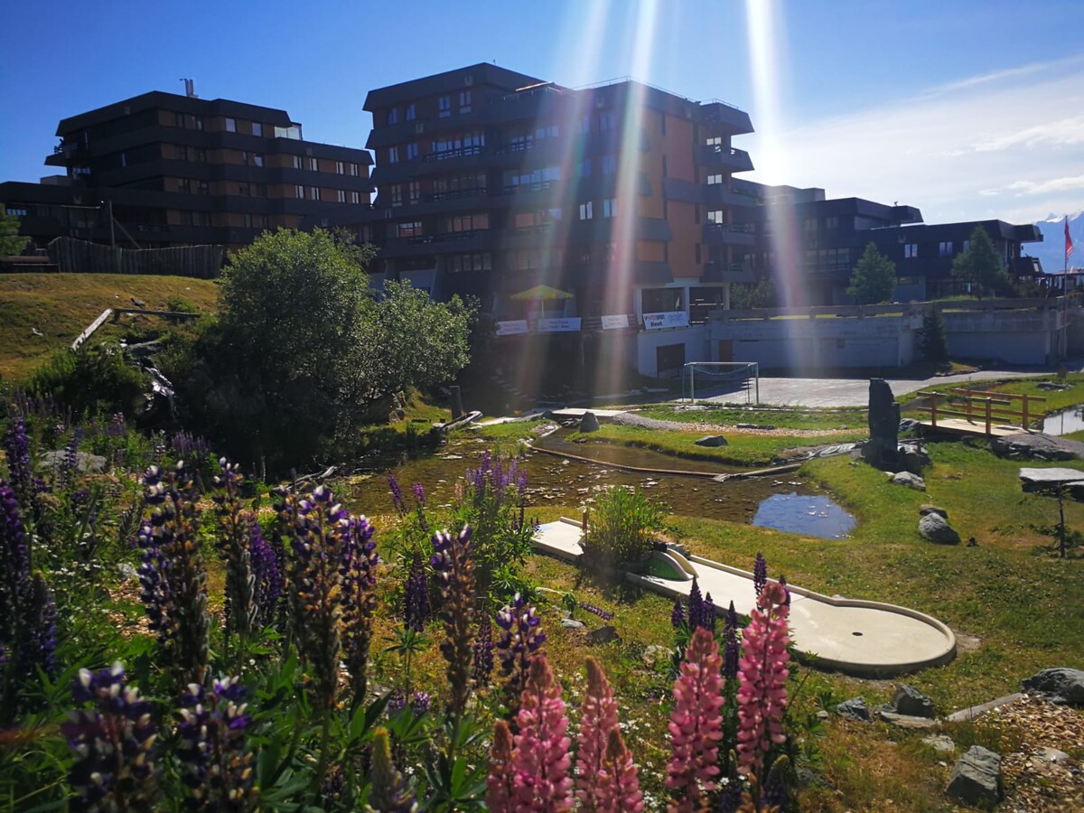 Colorful flowers frame a serene outdoor area, leading toward a lodge-style building complex. A small pond reflects the sky, while a grassy expanse features recreational amenities, including open spaces for leisure activities. The scene conveys a tranquil and natural setting suitable for relaxation.