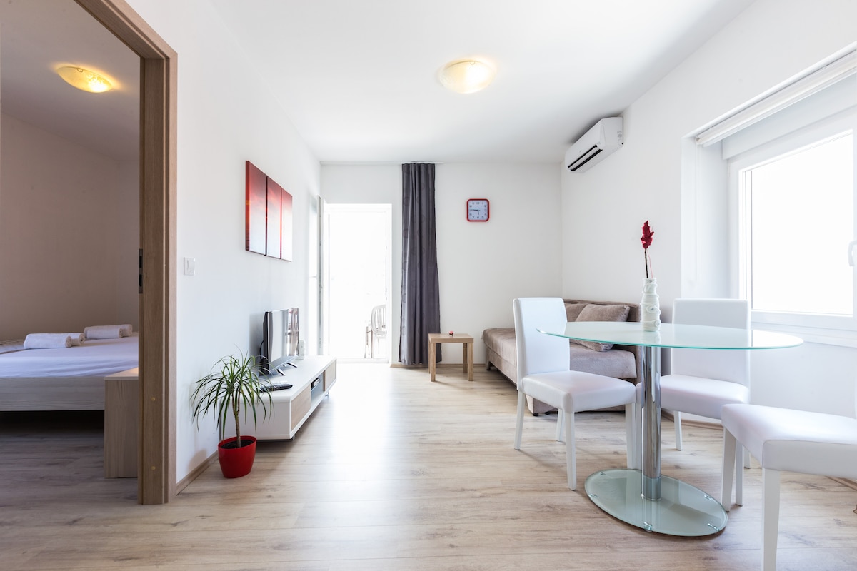 A bright living space features a glass dining table with white chairs, complemented by a comfortable sofa and a television on a sleek unit. Natural light floods in through the large windows, creating an inviting atmosphere. A small plant adds a touch of greenery.
