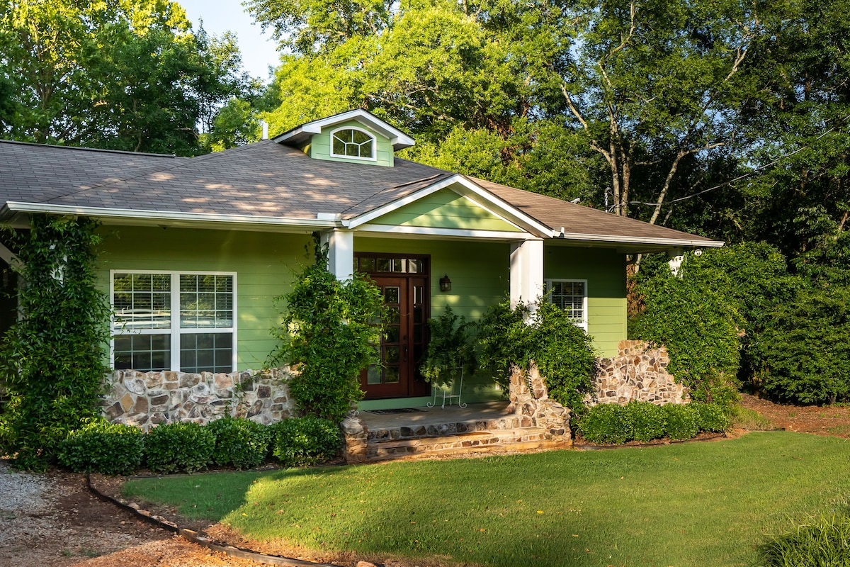A charming cottage exterior is framed by lush greenery and natural stone accents. The front porch features white columns and a welcoming entrance, with large windows allowing natural light to enhance the inviting facade.