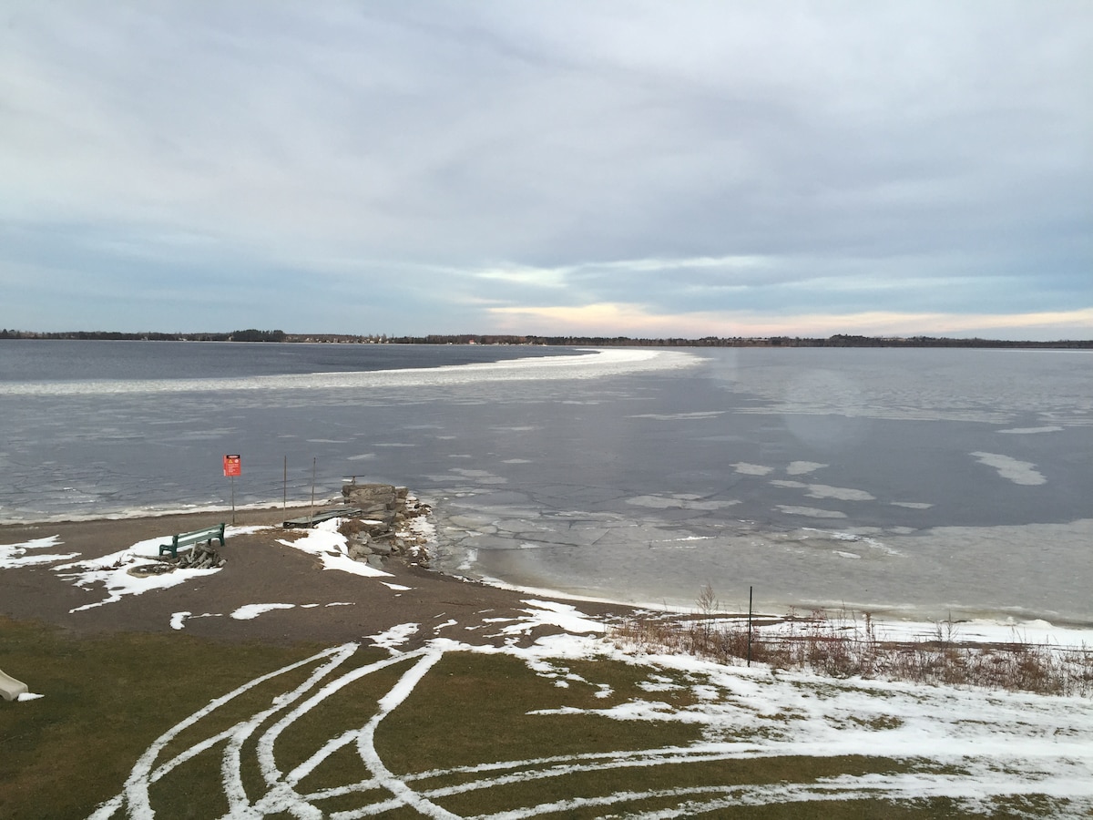 The serene view of Lac-Saint-Jean is showcased, with a frozen surface reflecting shades of grey and blue. A snow-covered shoreline is visible, with a hint of a pathway leading towards the water. The distant horizon features a soft light, indicating the transition from day to evening.