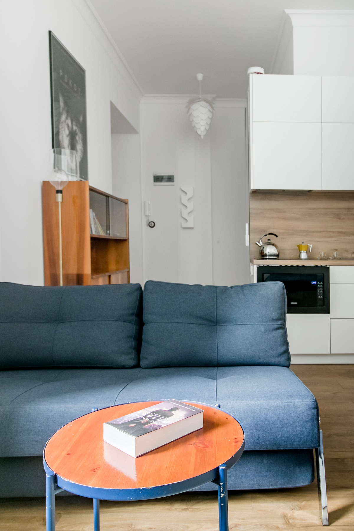 A cozy seating area is highlighted by a blue designer sofa, paired with a round wooden table. A book rests on the table's surface. The background reveals a modern kitchen with white cabinetry and a microwave, contributing to the tidy and functional atmosphere.