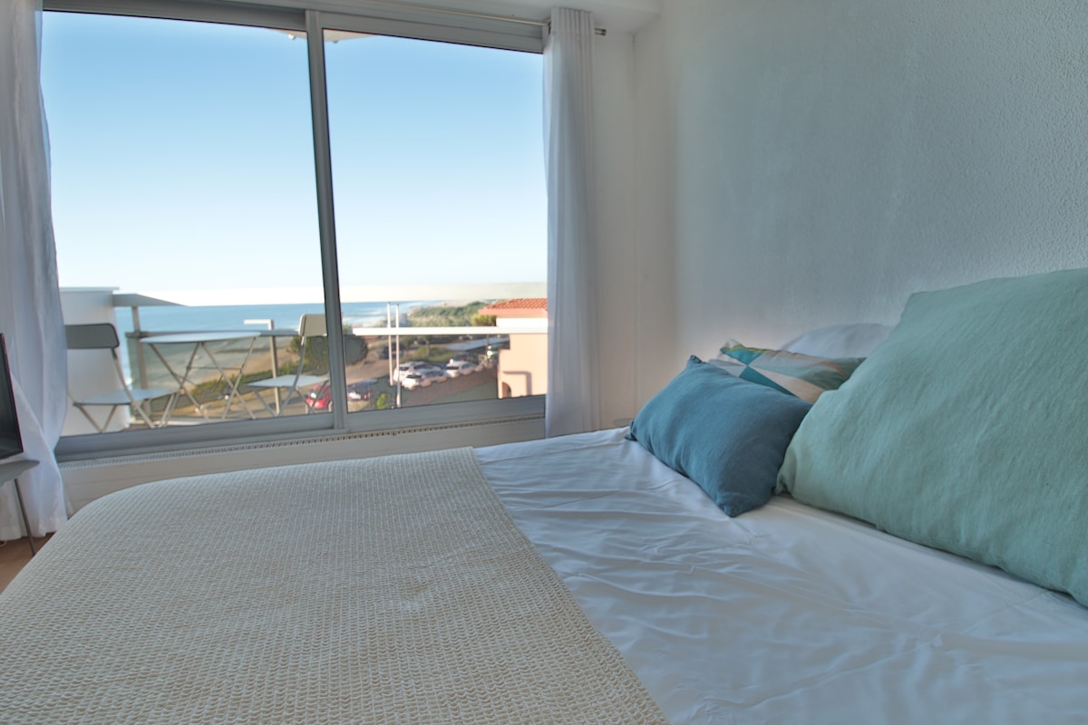 A spacious sleeping area features a bed with soft pillows and a light blanket, illuminated by natural light. A large window offers a scenic view of the ocean and distant coastline, while a small balcony is visible in the background.