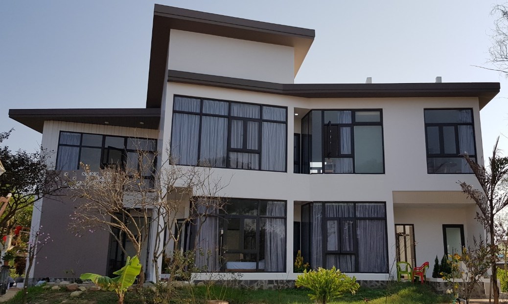 A modern building presents a sleek architectural design, featuring large windows that invite natural light. The exterior is adorned with minimal landscaping, including small trees and green plants. The structure is layered, showcasing a unique roofline that adds visual interest.