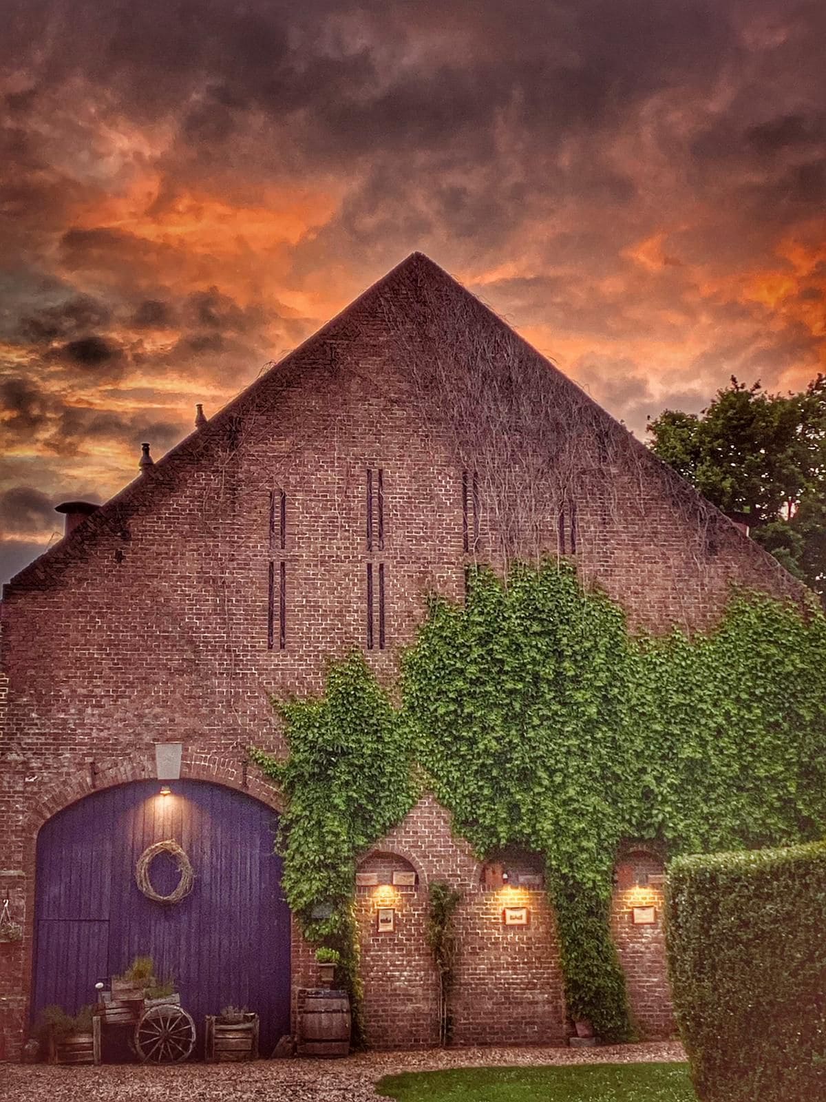 A historic building features a thatched roof and large arched entrance, partially covered by lush greenery. Soft glowing lights highlight the entryway, while the dramatic sky casts vibrant hues, creating an inviting setting against the brick structure.