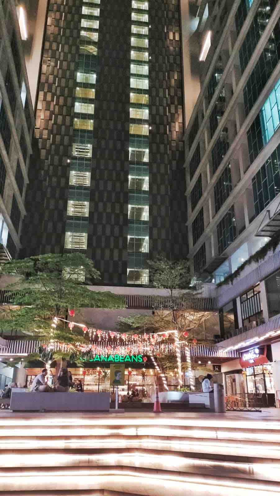 An urban plaza is framed by tall buildings, with a landscaped area illuminated by decorative lights. Outdoor seating is arranged in front of a café, creating a welcoming space for relaxation. The contemporary architecture showcases a mix of glass and patterned facades.