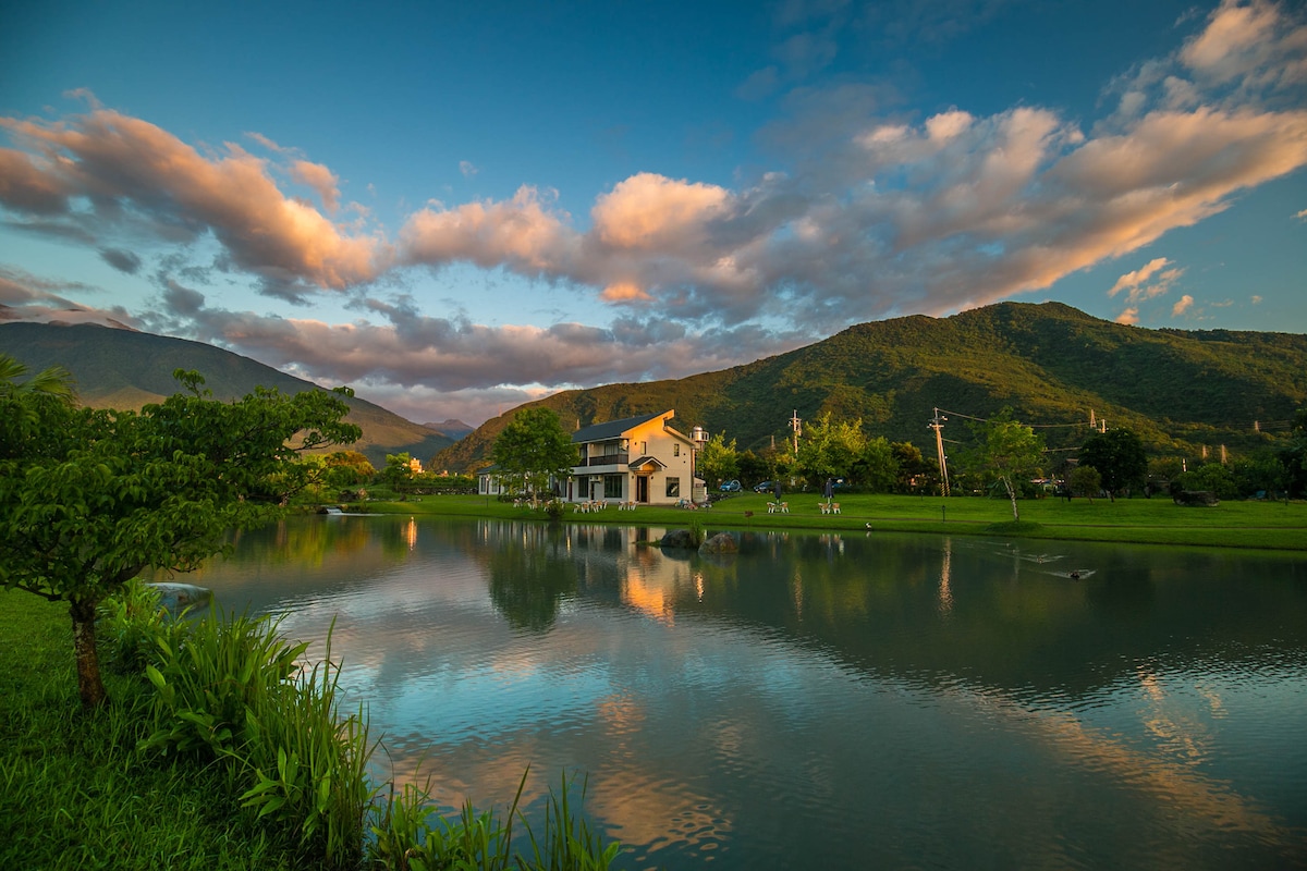 A tranquil landscape features a serene lake reflecting lush green surroundings and a charming house. Soft clouds drift above the distant mountains, while the sky displays gentle hues of blue and orange. The inviting lawn is bordered by vibrant greenery, enhancing the peaceful setting.