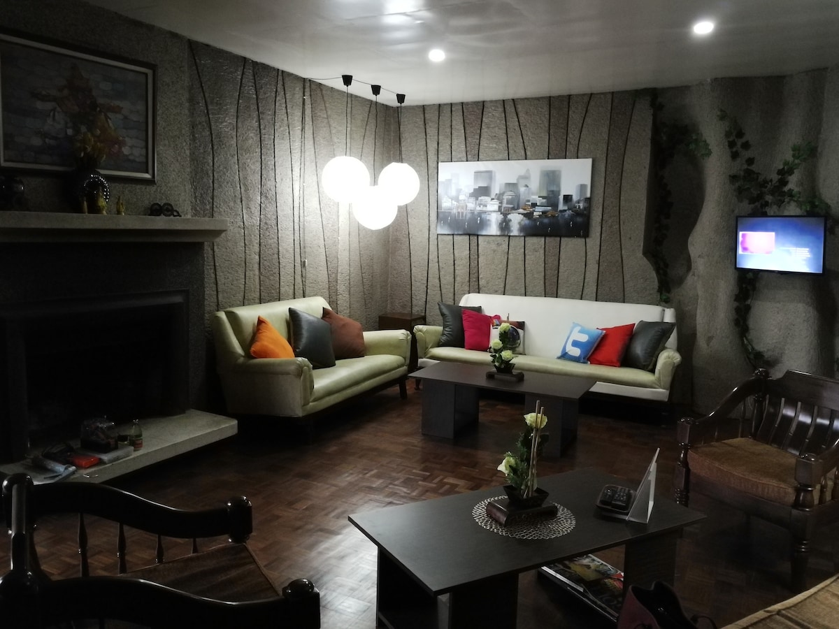 The spacious living room features a stone wall with artistic textures and a cozy seating arrangement. Two sofas, accented with colorful cushions, and a wooden chair are arranged around a central coffee table. A flat-screen TV is mounted on the wall, complemented by ambient lighting.