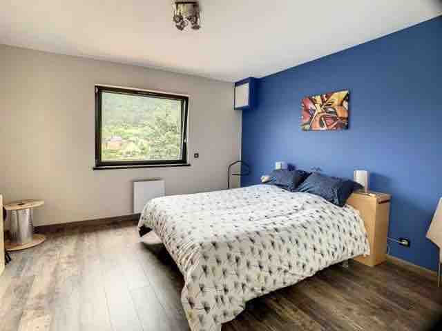 A serene bedroom features a spacious bed with a patterned duvet. A vibrant blue accent wall is complemented by a piece of abstract artwork. A large window invites natural light and offers a view of the surrounding landscape. Simple nightstands are positioned on either side.