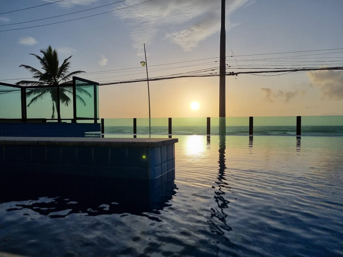 An infinity pool extends towards the sea, its water reflecting the warm hues of the sunset. In the background, a palm tree stands silhouetted against the horizon, while utility poles mark the edge of the view.