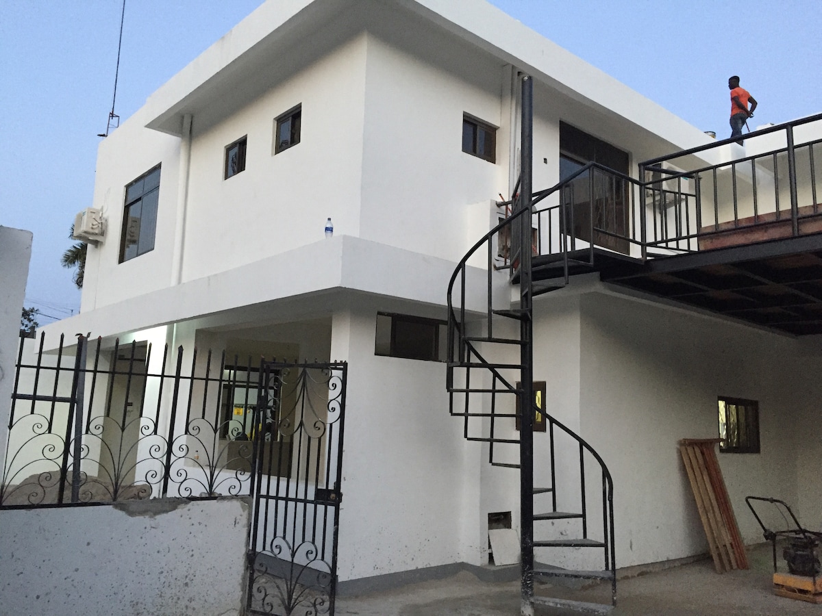 An exterior view of the house features a clean, modern design with a white facade and windows framed in dark materials. A spiral staircase leads to an upper level, and a gated entrance is visible in the foreground. The quiet neighborhood setting is highlighted.