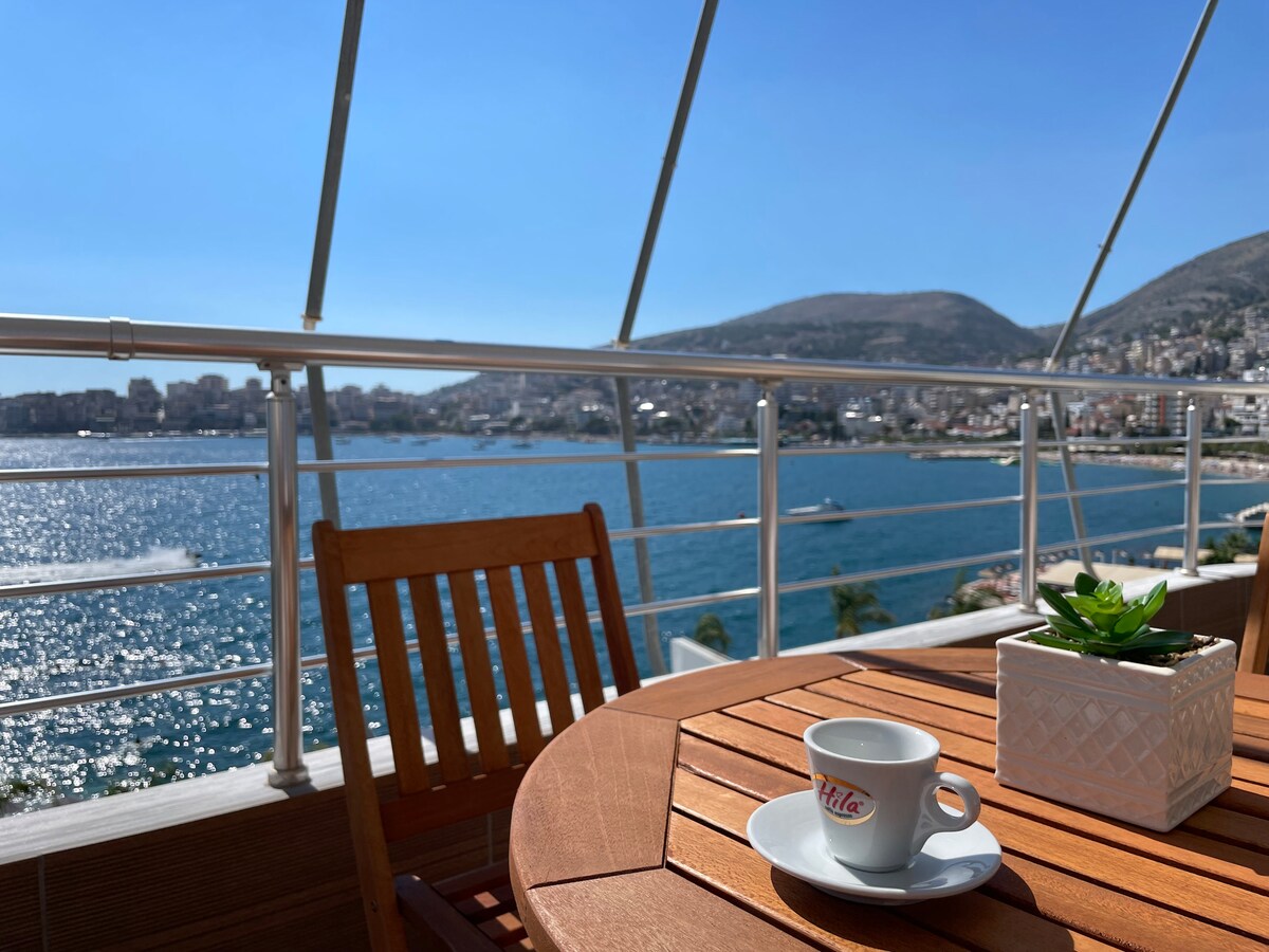 A balcony table is set with a white coffee cup and a small potted plant. The vibrant sea view is complemented by distant hills and a bustling shoreline, showcasing a mix of watercraft and coastal buildings under clear blue skies.