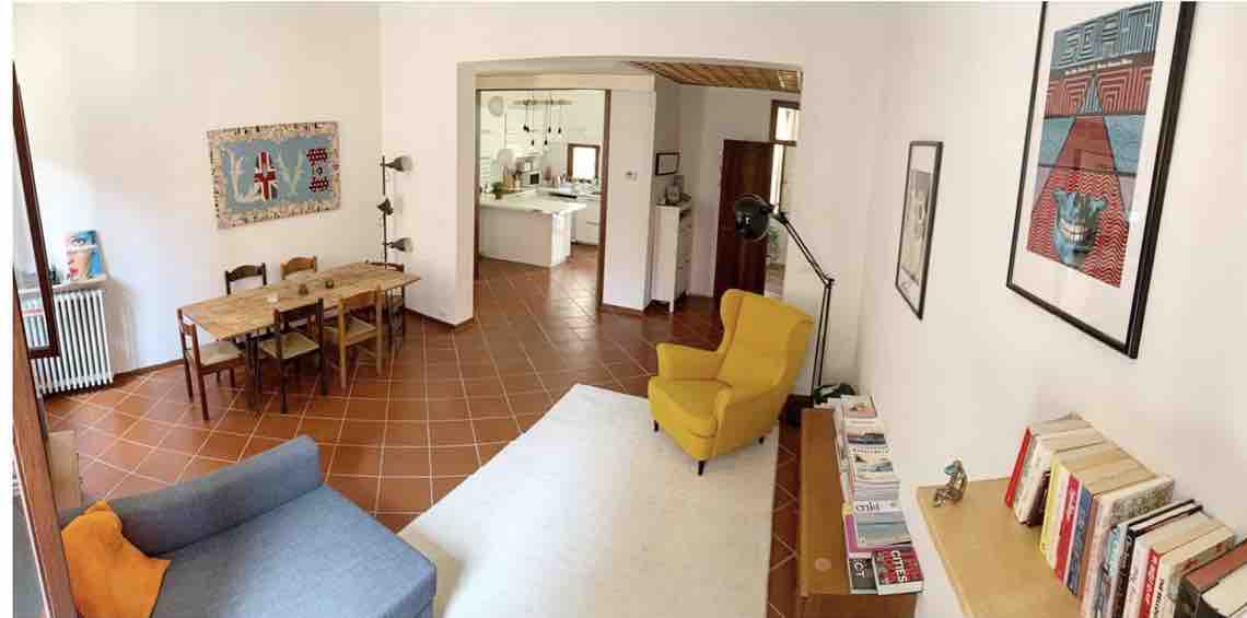 An airy living space is depicted, featuring a blue sofa and a vibrant yellow armchair. A rustic wooden dining table with chairs is seen in the background. The space is enhanced by decorative art pieces on the walls and warm terracotta tile flooring.