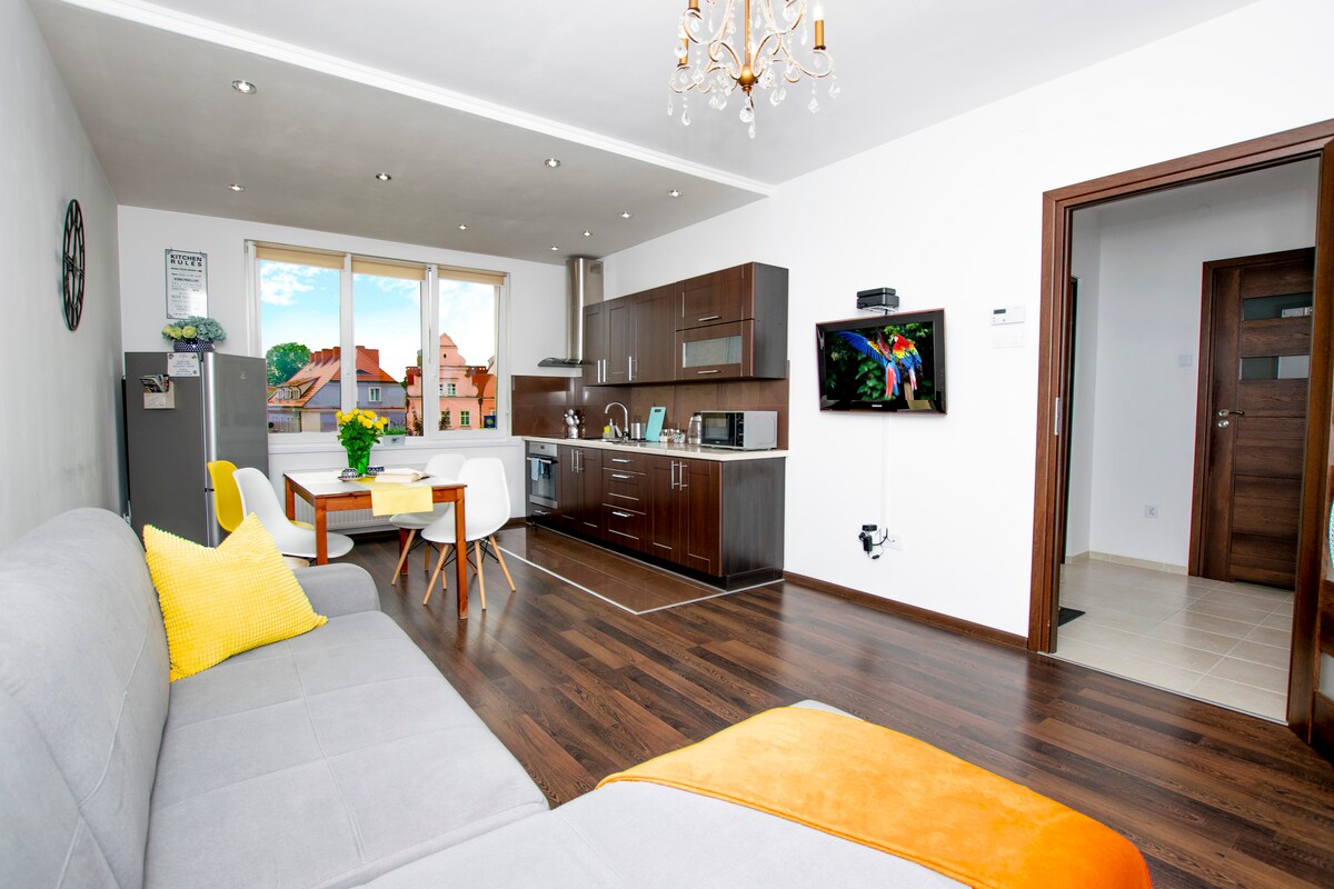 The living area features a comfortable gray sofa, paired with a bright yellow pillow. A wooden dining table with two chairs is positioned near large windows that offer a view of the surrounding architecture. An LCD TV is mounted on the wall, with modern cabinets visible in the kitchen area.