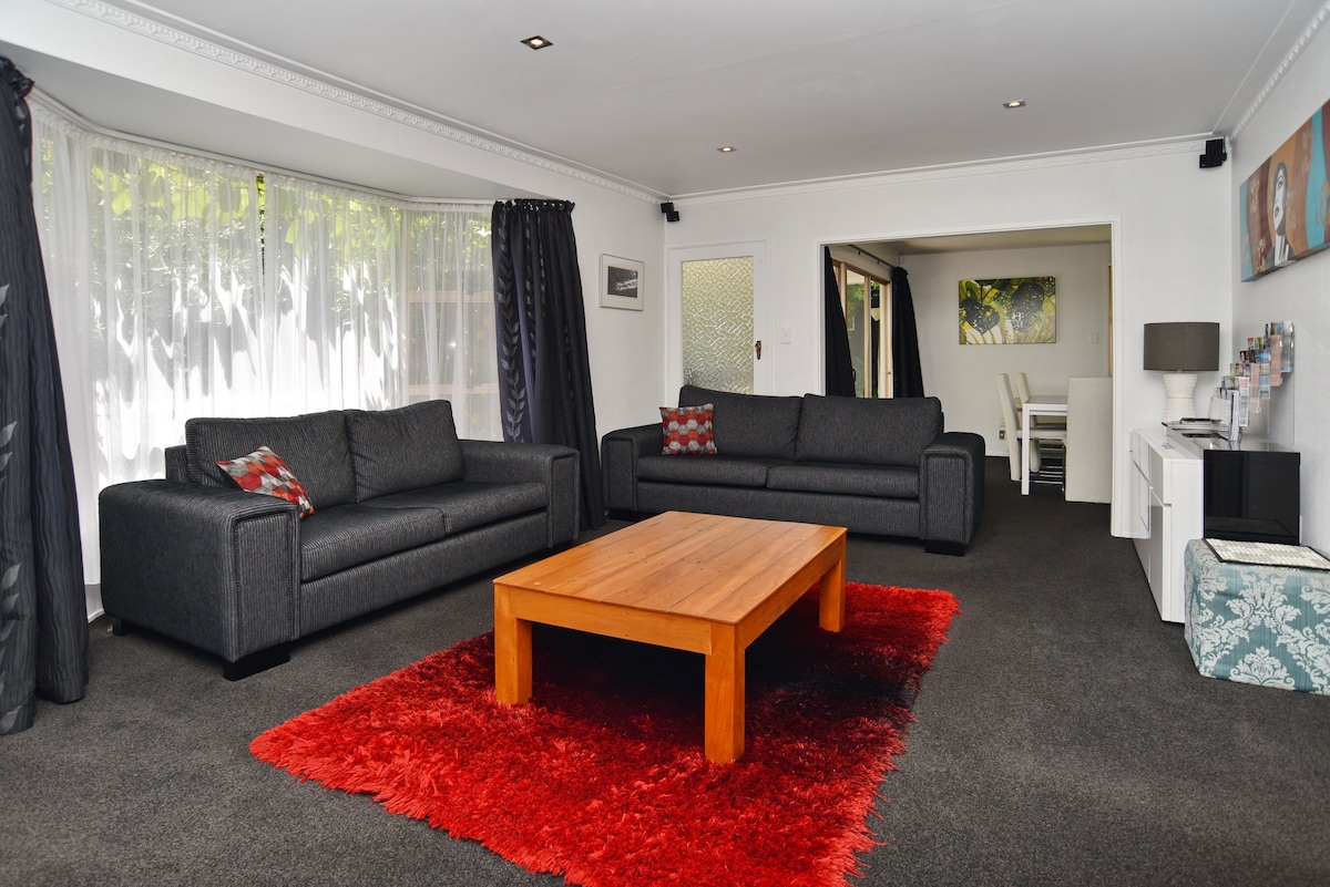 A spacious living area is presented with two dark grey sofas positioned around a wooden coffee table atop a vibrant red rug. Soft natural light enters through large windows, draped with sheer white curtains. A dining area is visible in the background, offering a bright and inviting atmosphere.