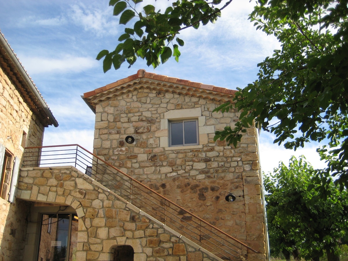 The exterior of the stone building is presented, featuring a staircase leading to an upper level. The upper window is visible, framed by a neutral stone façade. Trees and greenery are seen surrounding the structure, contributing to a natural setting.