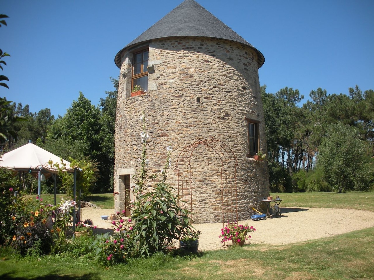 The renovated windmill features a circular stone exterior, enhanced by a conical roof. Surrounding vegetation and flowers are visible, contributing to a serene outdoor space. A gravel pathway leads to the entrance, framed by greenery and a tent structure nearby.
