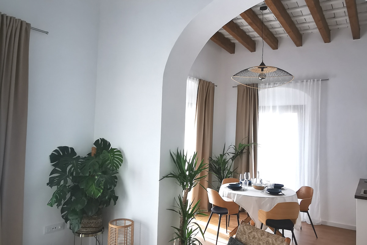 A bright dining area is displayed, featuring a round table set for four. Wooden beams span the ceiling, enhancing the open atmosphere. Potted plants are strategically placed, and large windows allow natural light to fill the space, complemented by soft curtains.