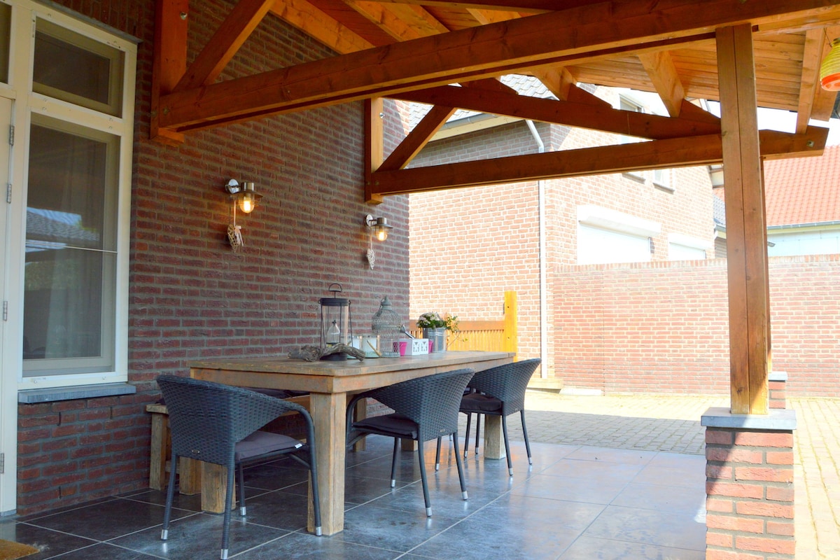 A covered terrace area features a wooden dining table surrounded by wicker chairs. Soft lighting is provided by wall-mounted lanterns, and the flooring consists of large tiles. The space offers a view of the courtyard area, illuminated by natural light.