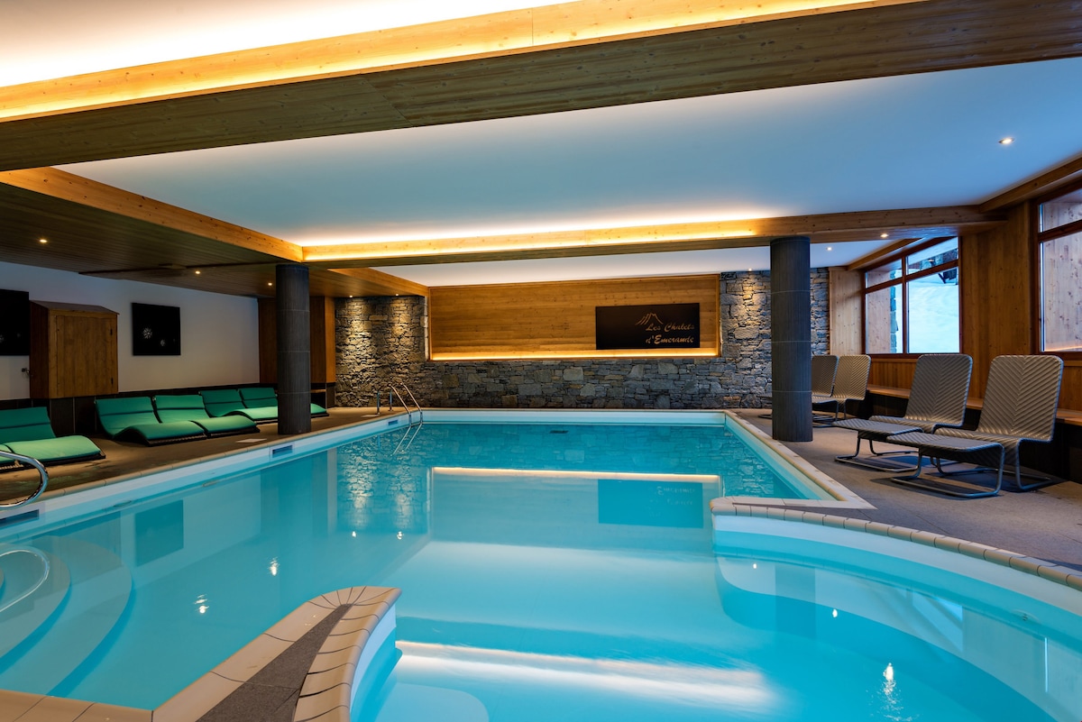 An indoor pool area features smooth turquoise water under warm lighting. Comfortable loungers are positioned around the pool, while stone and wood accents create a serene atmosphere. Large windows allow natural light to fill the space, enhancing the tranquil environment.