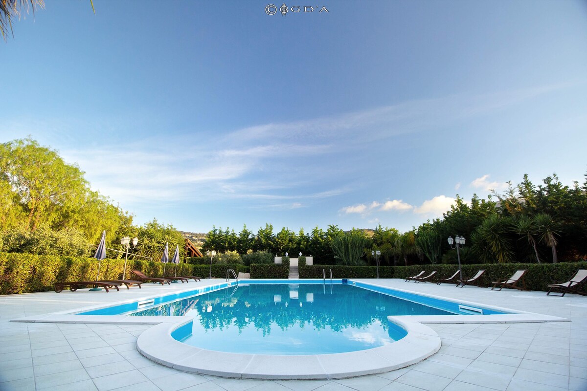 A large outdoor swimming pool is surrounded by lush greenery and neatly arranged sun loungers. The pool features a shallow section and is set on a spacious tiled patio, offering a serene environment for relaxation.
